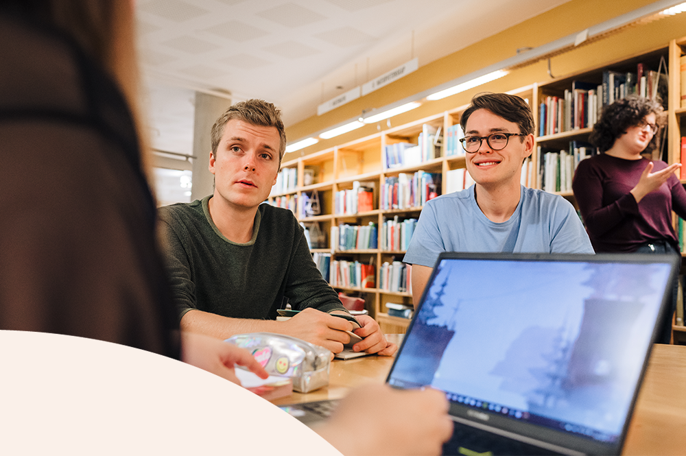 Foto som föreställer två studenter vid varsin dator, pratandes med någon bakom kameran